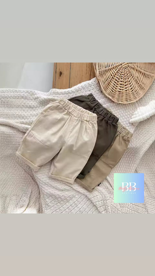 Three pairs of shorts in beige, brown, and green on a textured surface with a wicker basket in the background.