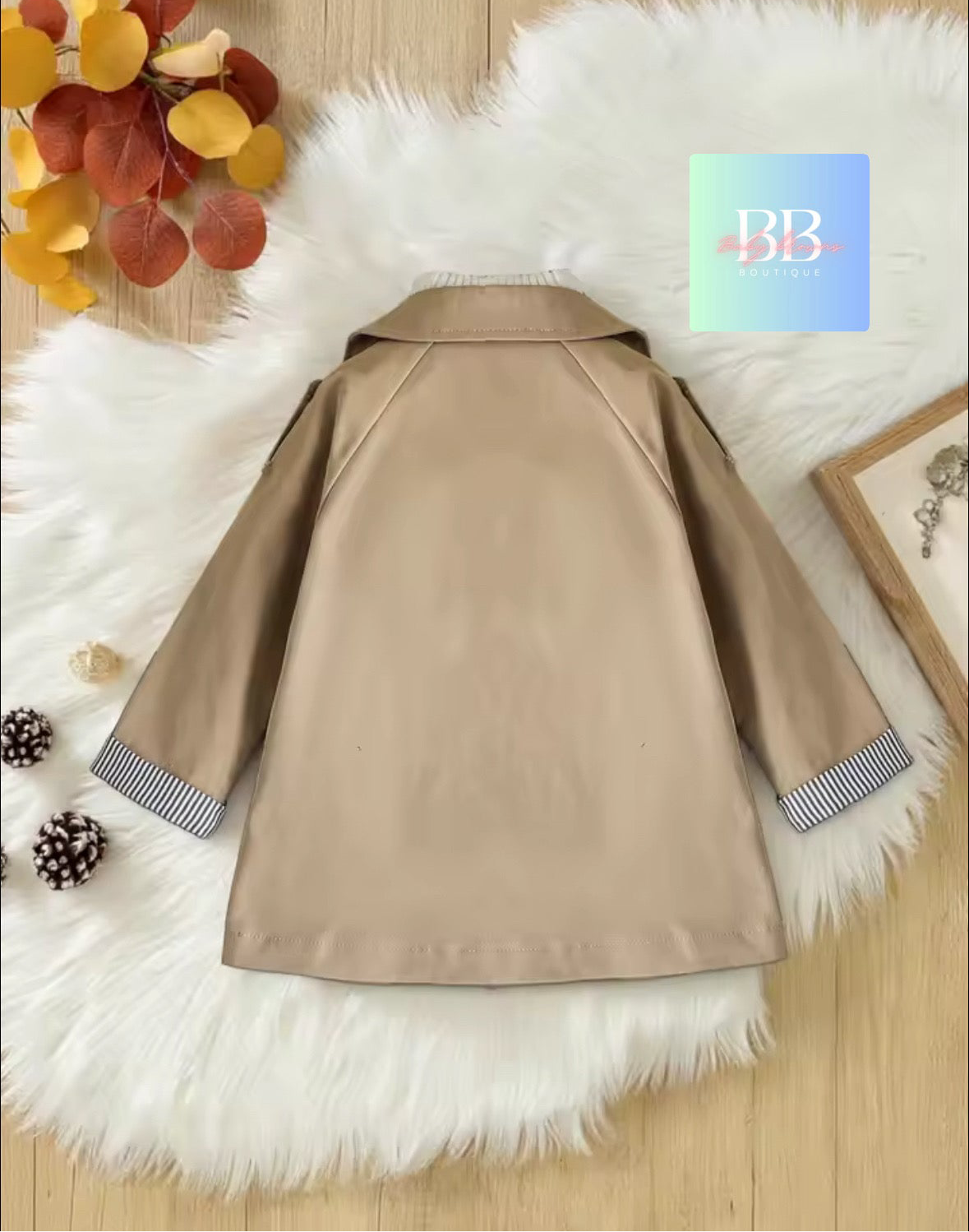 Beige coat with fur trim on a fluffy white surface, surrounded by autumn leaves and pine cones.