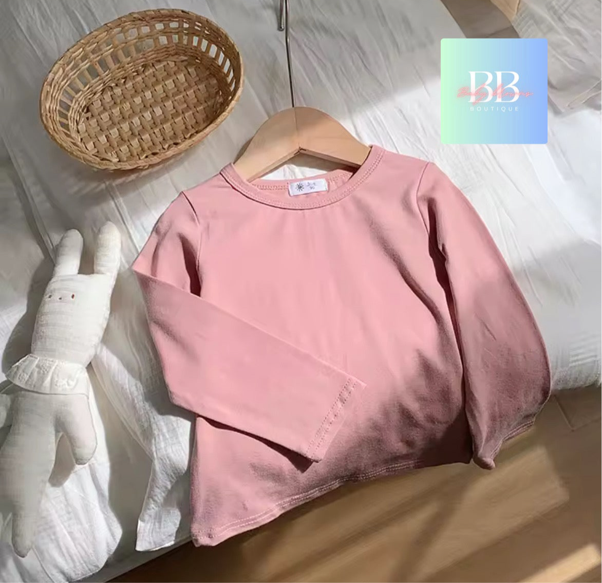 Pink sweatshirt on a wooden hanger with a white glove and woven basket on a light wooden floor.