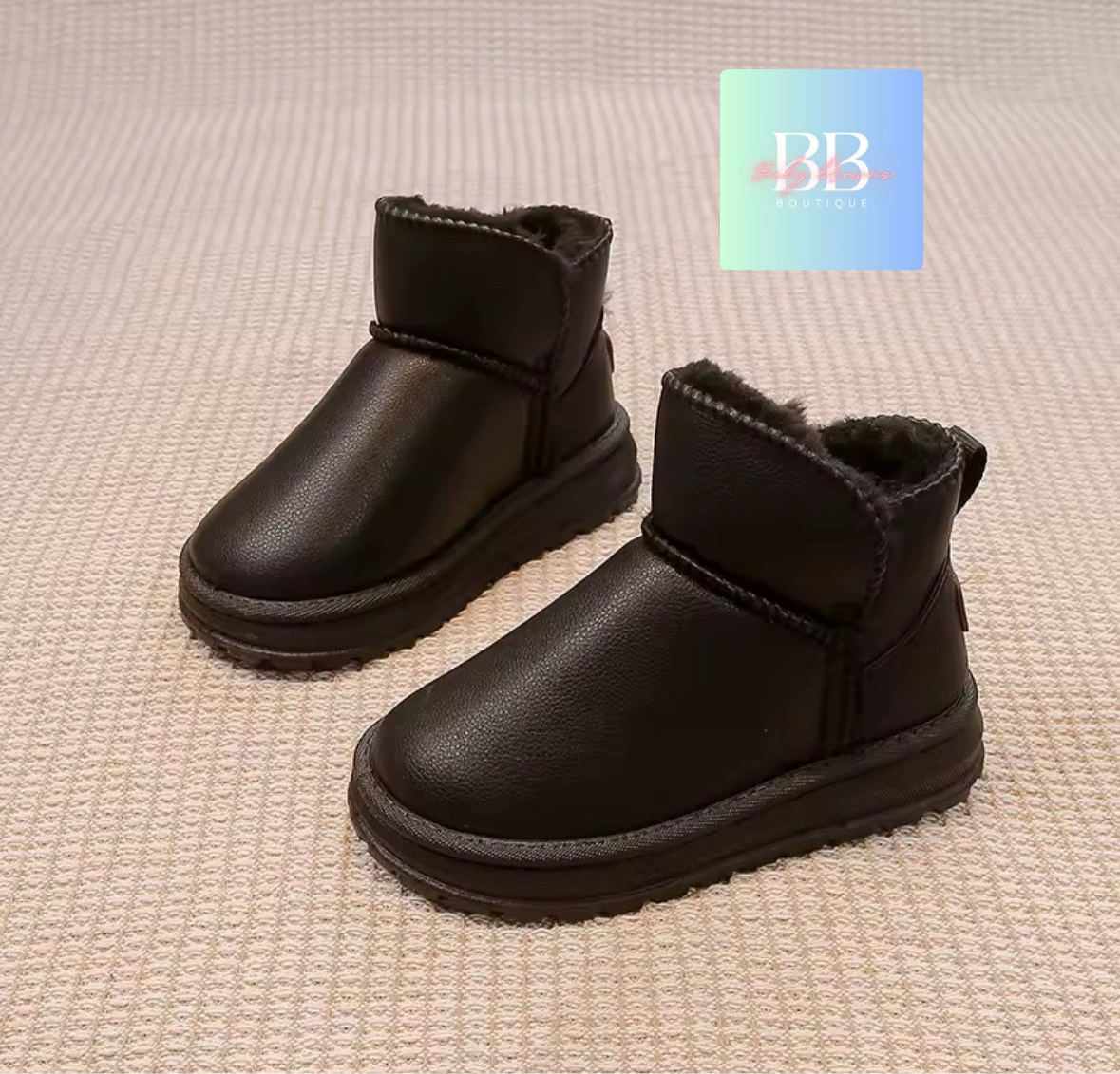 Pair of black children's boots on a textured beige surface with a brand logo in the corner.
