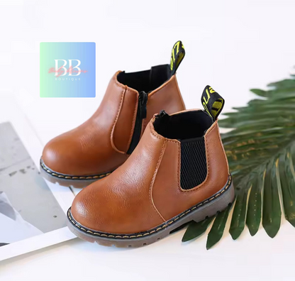 Pair of brown children's boots with black soles on a white surface with green leaves.