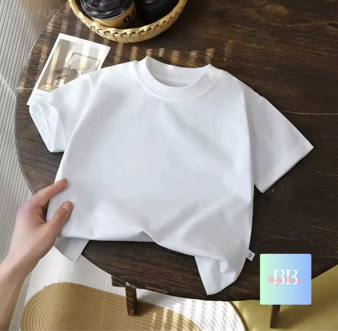 White t-shirt on a wooden table with a hand holding it, and a coffee cup in the background.