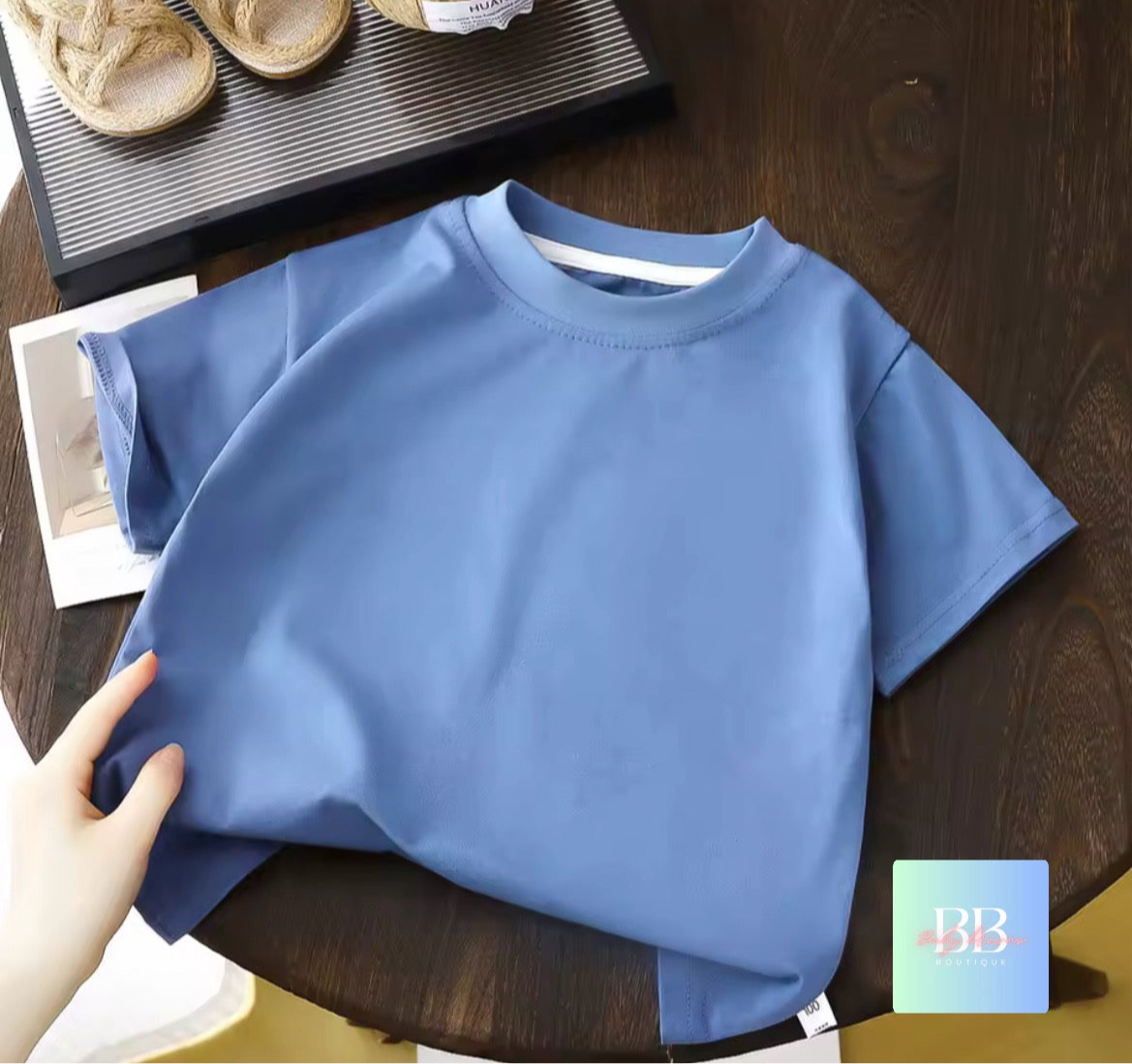 Blue shirt on a wooden table with a hand holding it, and a logo in the corner.