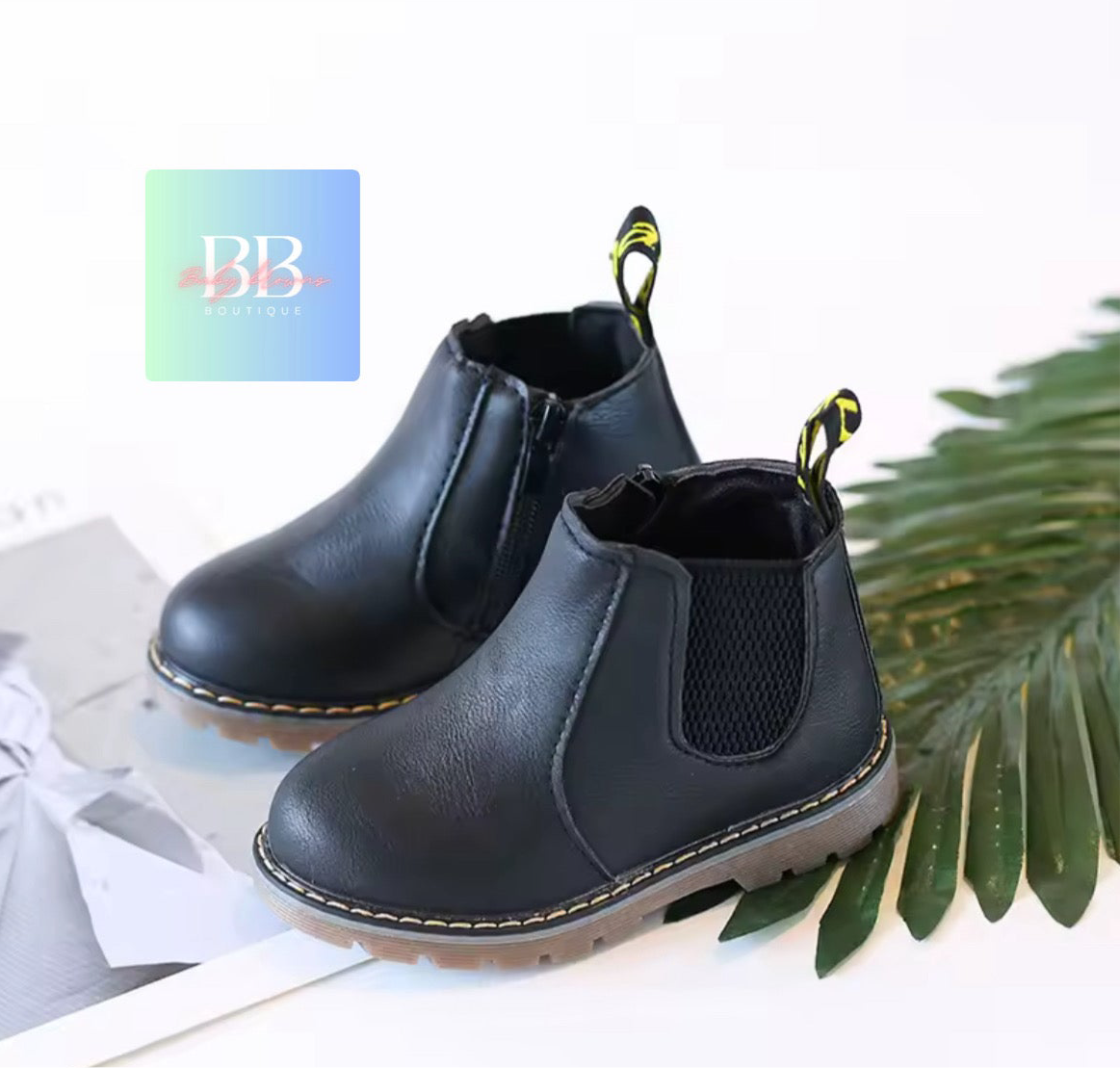 Black boots with yellow accents on a white surface with a green leafy plant.