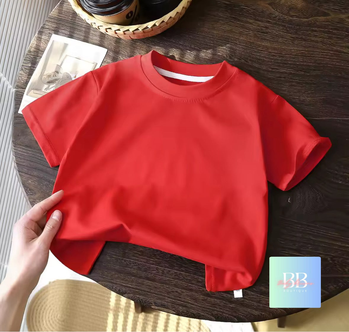 Red shirt on a wooden table with a hand holding it, and a small blue square with 'BB' logo in the corner.