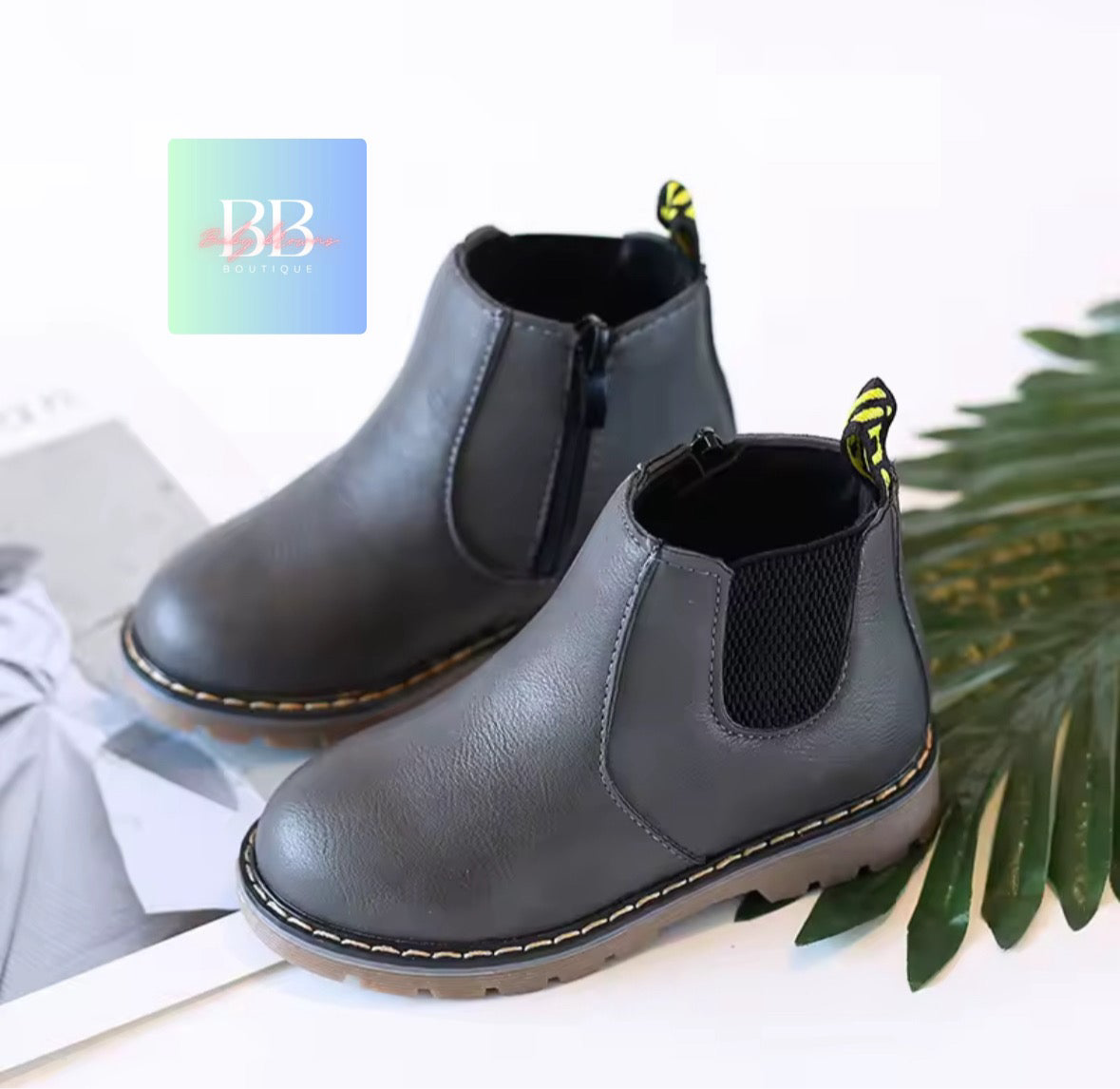 Pair of gray children's boots with yellow stitching on a white surface with green leaves.