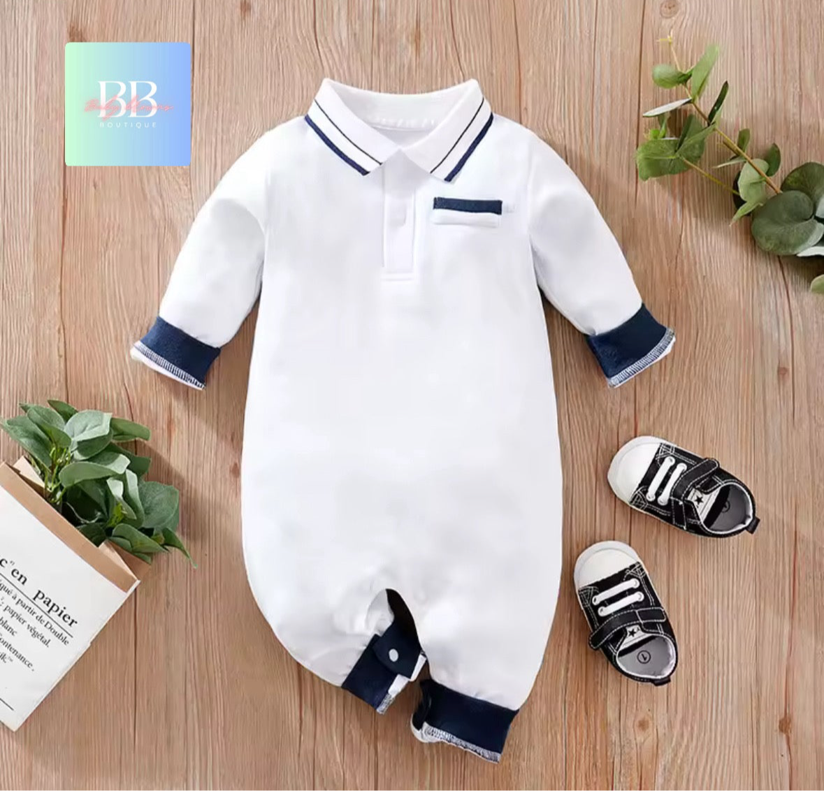 White baby romper with navy trim on a wooden surface, next to black and white shoes.
