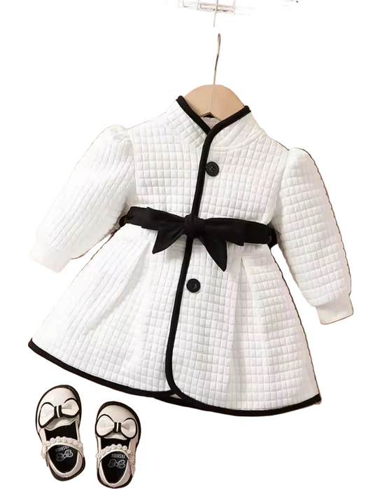 White quilted dress with black buttons and belt on a wooden surface, accompanied by black shoes.