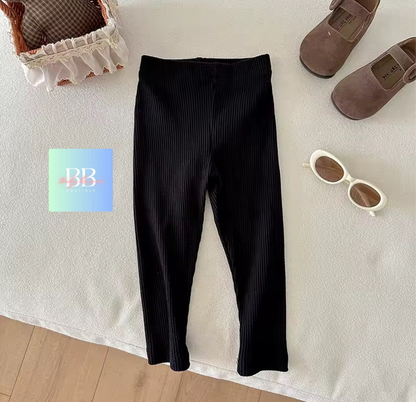 Black pants on a white surface with a basket, shoes, and sunglasses nearby.