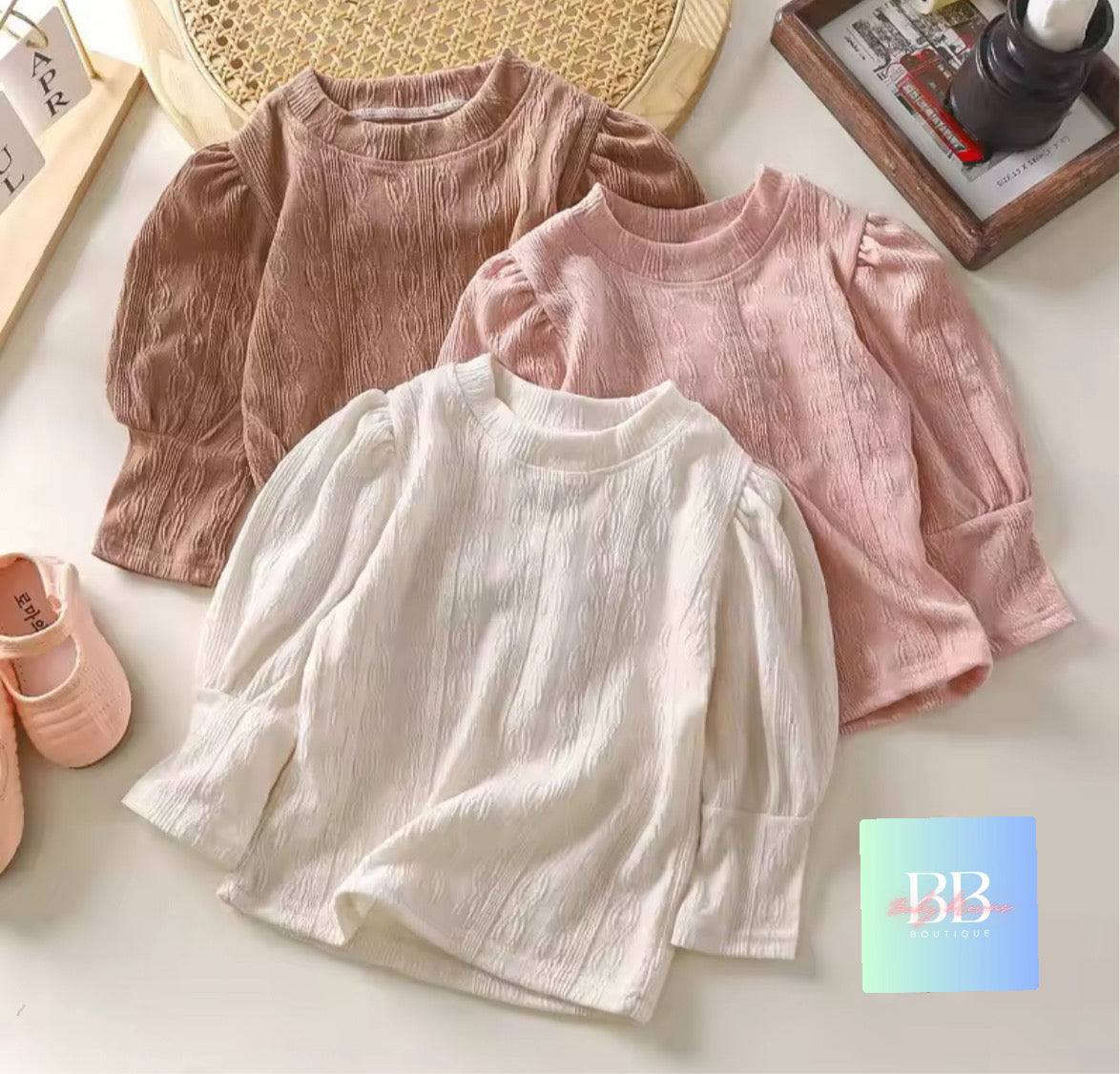 Three children's shirts in pink, white, and beige on a light surface with a BB logo in the corner.