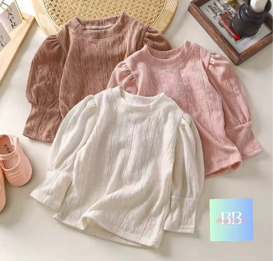 Three children's shirts in pink, white, and beige on a light surface with a BB logo in the corner.