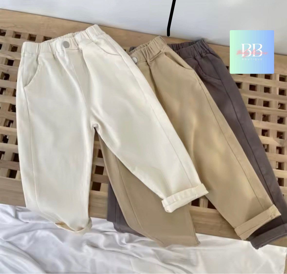 Three pairs of pants in beige, white, and brown on a wooden surface with a brand logo in the corner.