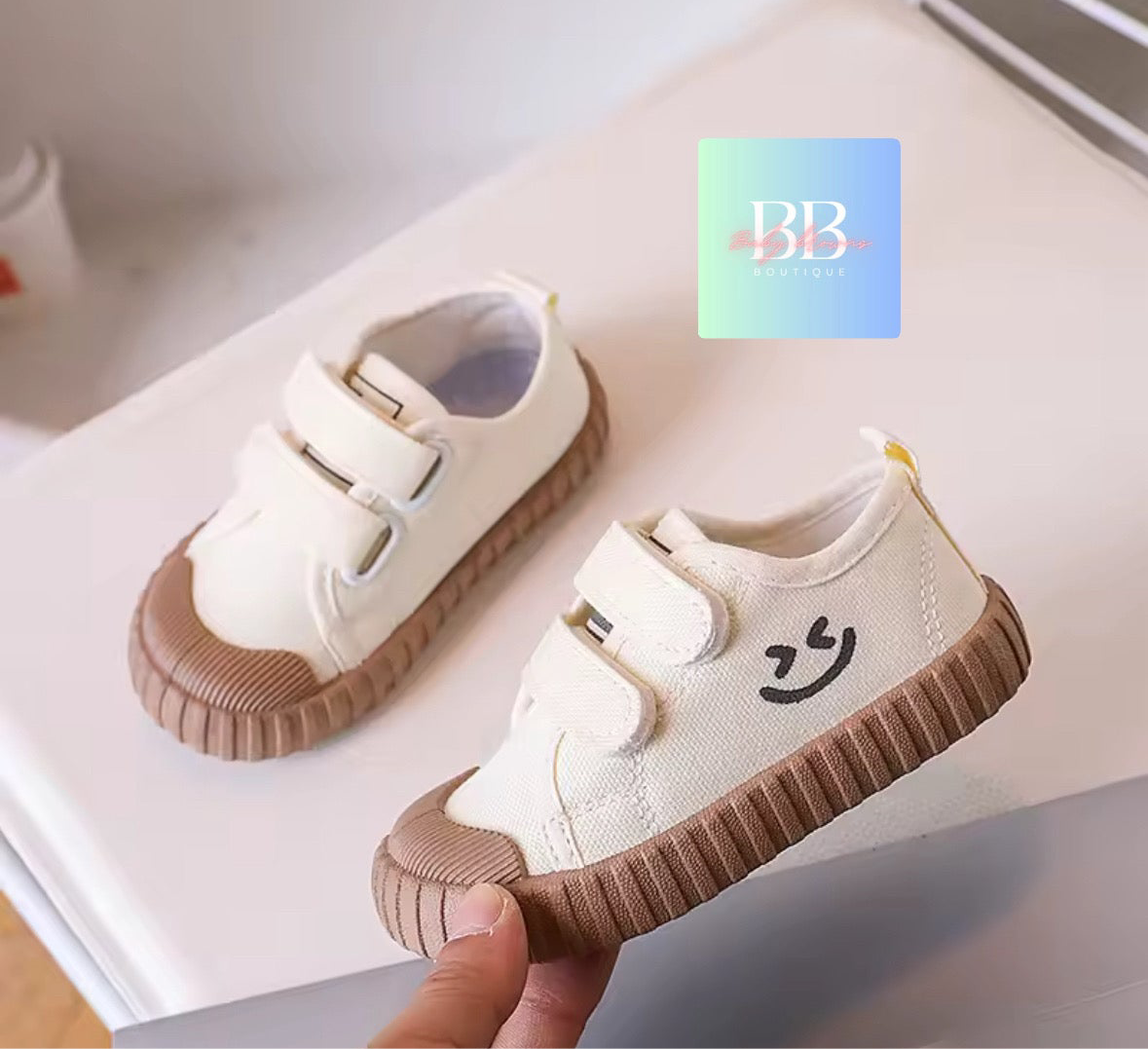 White children's shoes with brown soles and smiley face design, held by a hand.