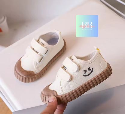 White children's shoes with brown soles and smiley face design, held by a hand.