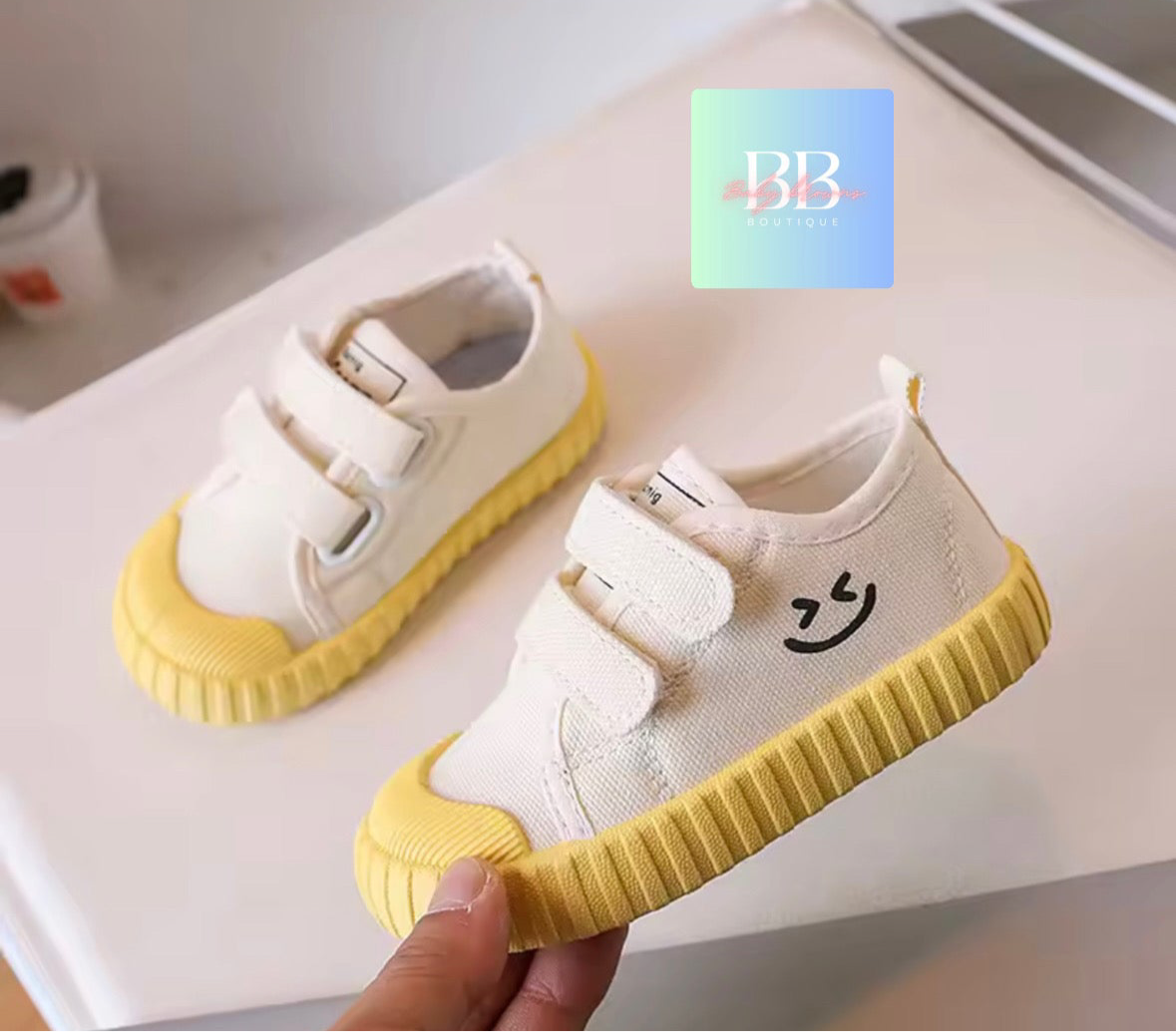 White children's shoes with yellow soles held by a hand on a light surface.