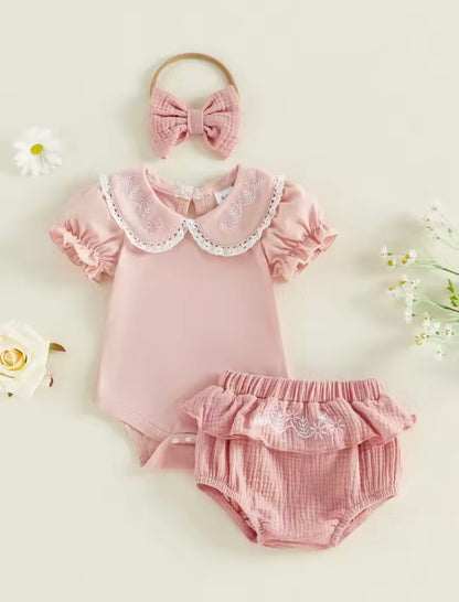 Pink baby outfit with ruffled collar, bloomers, and headband on a light background with flowers.