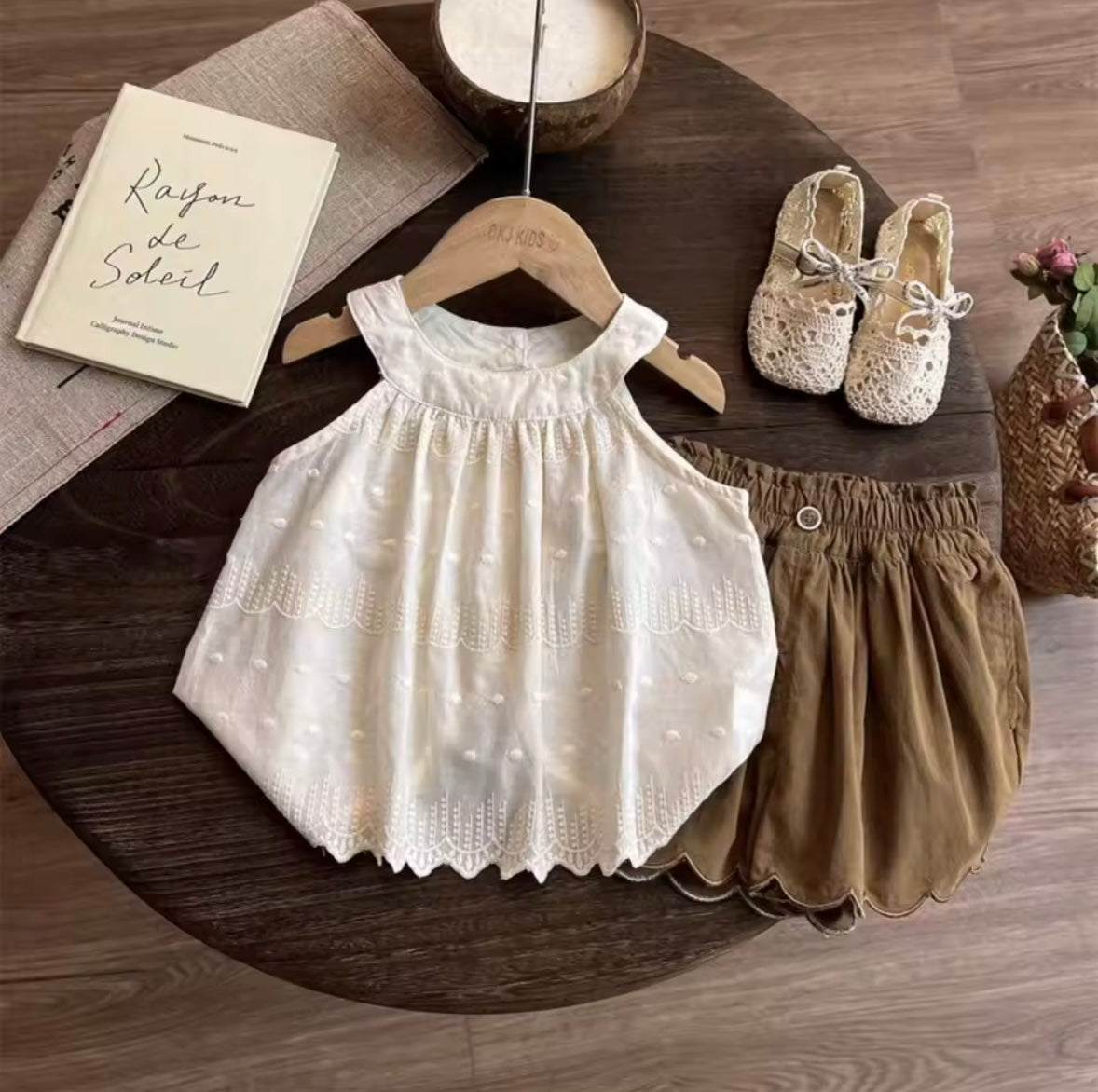Children's white dress and brown shorts on a wooden table with a book and shoes.