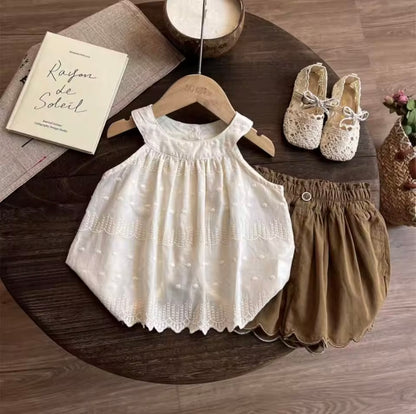 Children's white dress and brown shorts on a wooden table with a book and shoes.