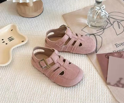 Pair of pink children's sandals on a textured surface with a book and decorative item.