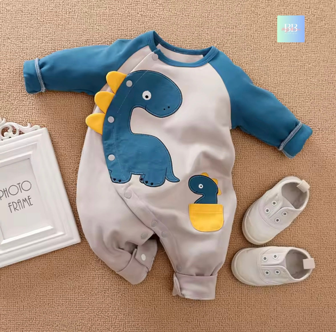 Baby romper with dinosaur design on a beige surface, next to a photo frame and baby shoes.
