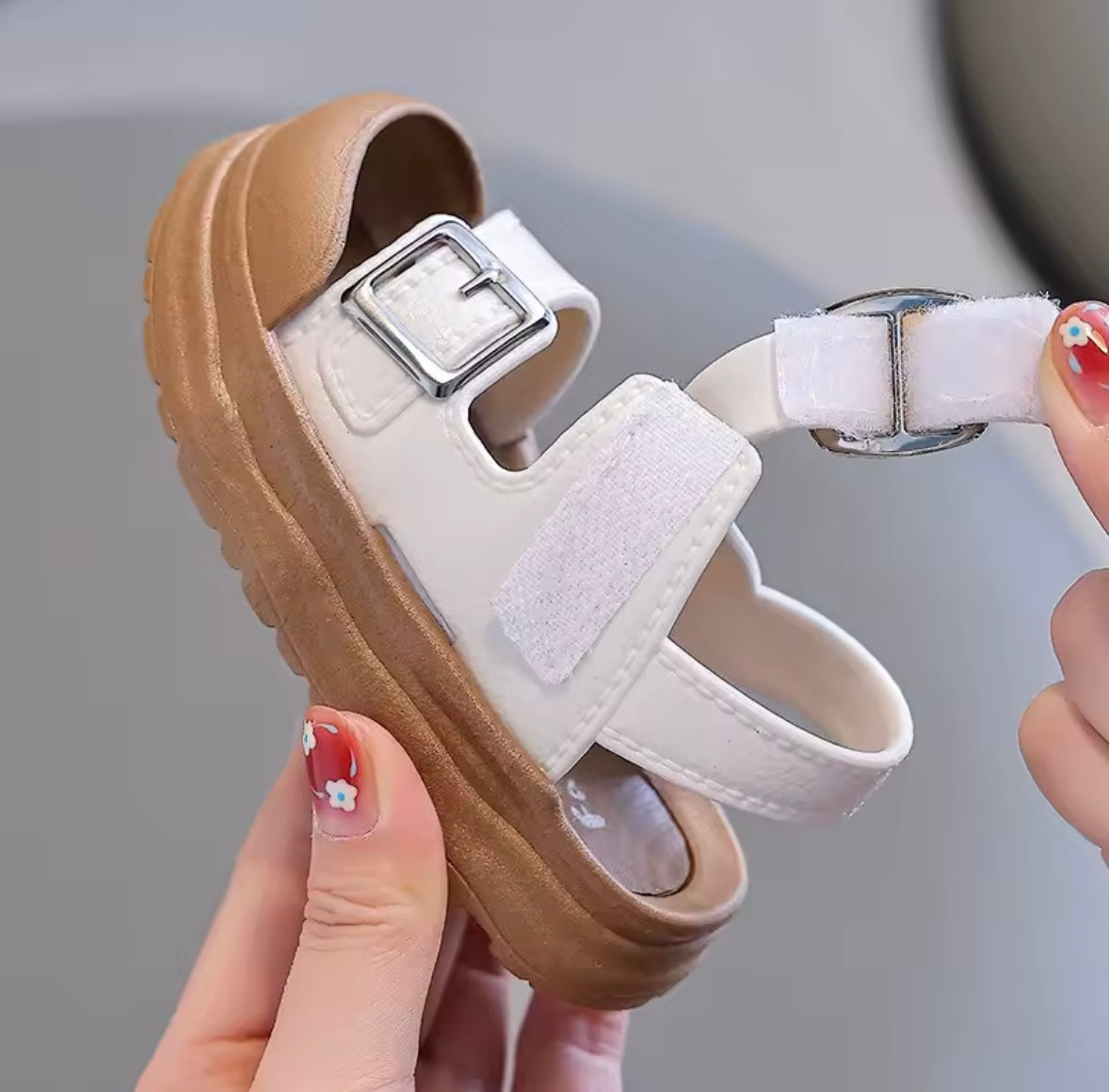 White sandal with brown sole held by a hand against a neutral background