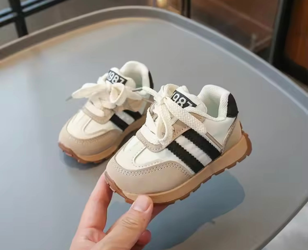 Pair of children's sneakers with black stripes held by a hand on a reflective surface.