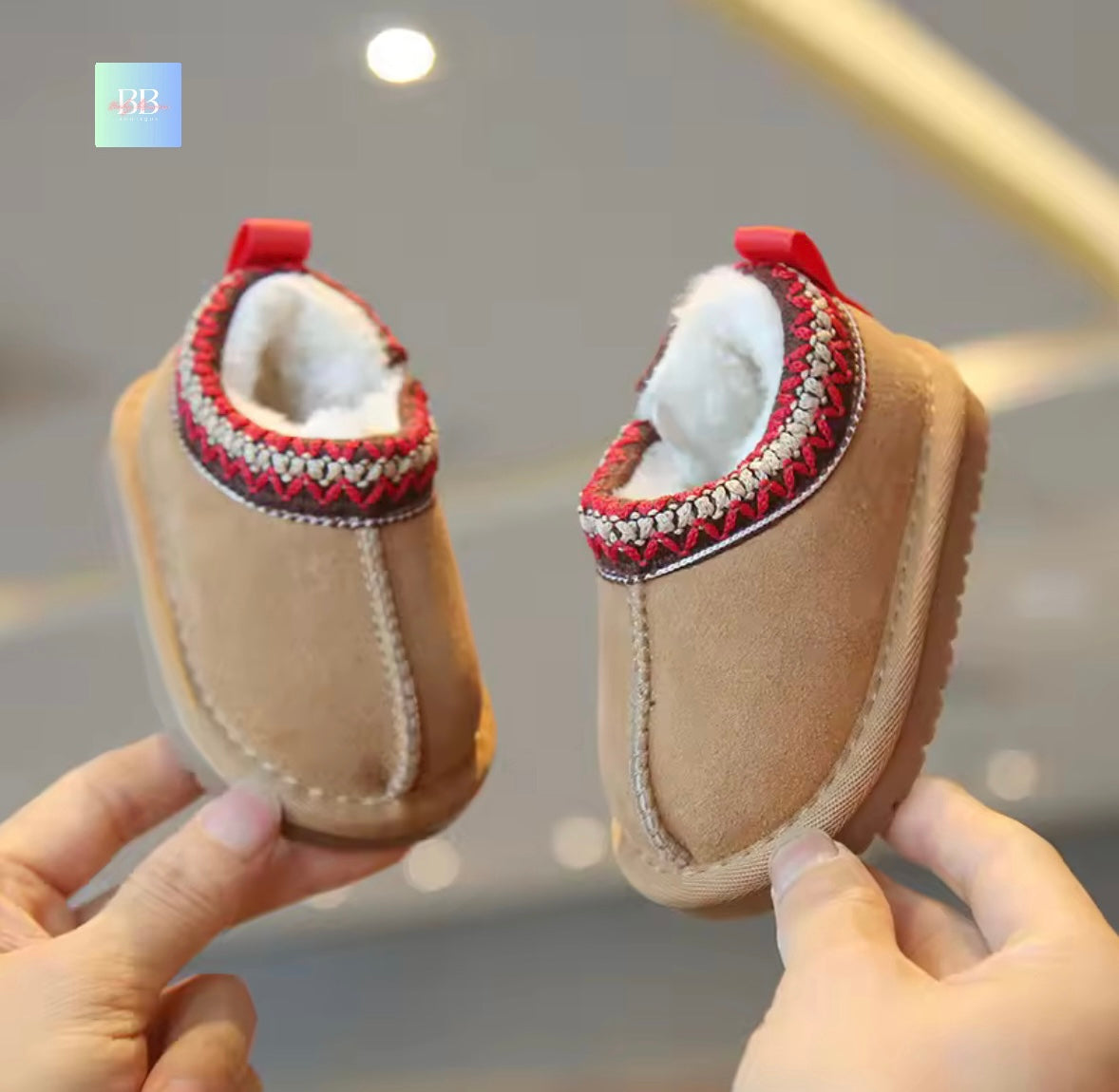 Pair of beige baby shoes with red and white accents held by hands against a blurred background