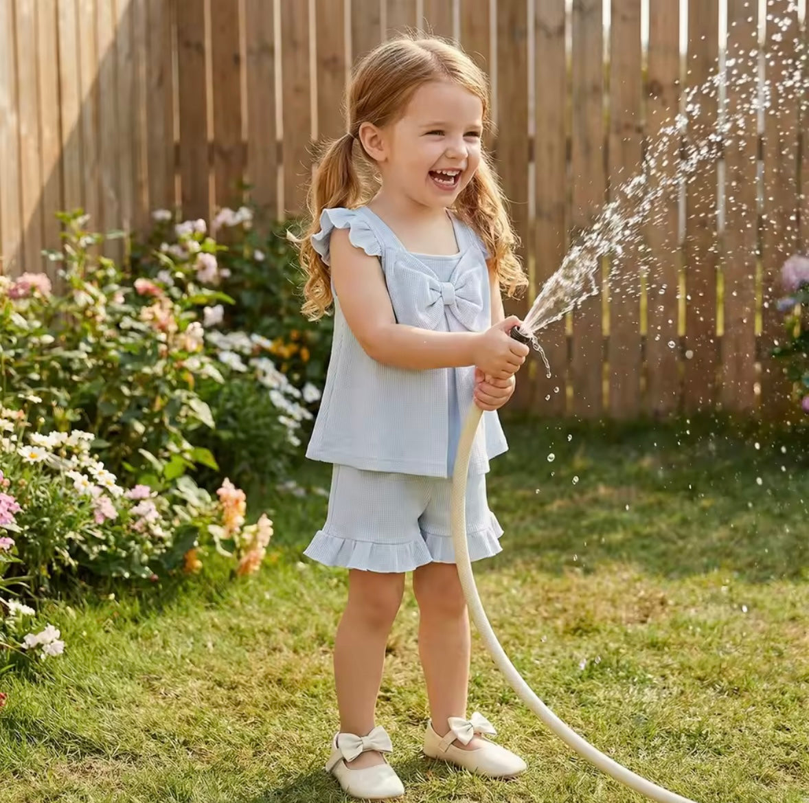 Young girl in a light blue dress playing with a garden hose in a backyard.
