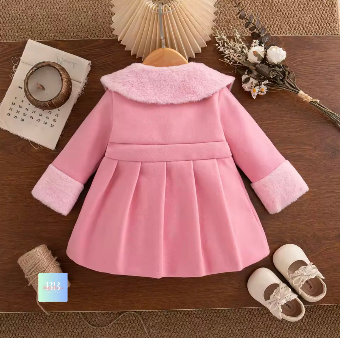 Pink children's coat with white trim on a wooden surface, surrounded by decorative items.