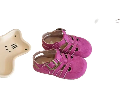 Purple children's sandals on a textured surface with a decorative plate and bottle in the background.
