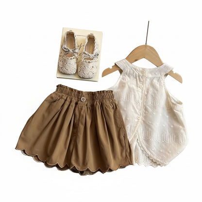 Children's clothing set including a white dress, brown shorts, and baby shoes on a wooden table.