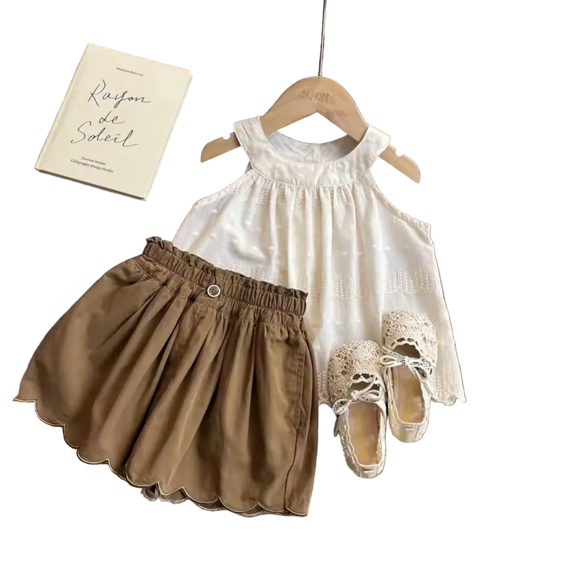 Children's white dress, brown skirt, and shoes on a wooden table with a book and fabric swatch.