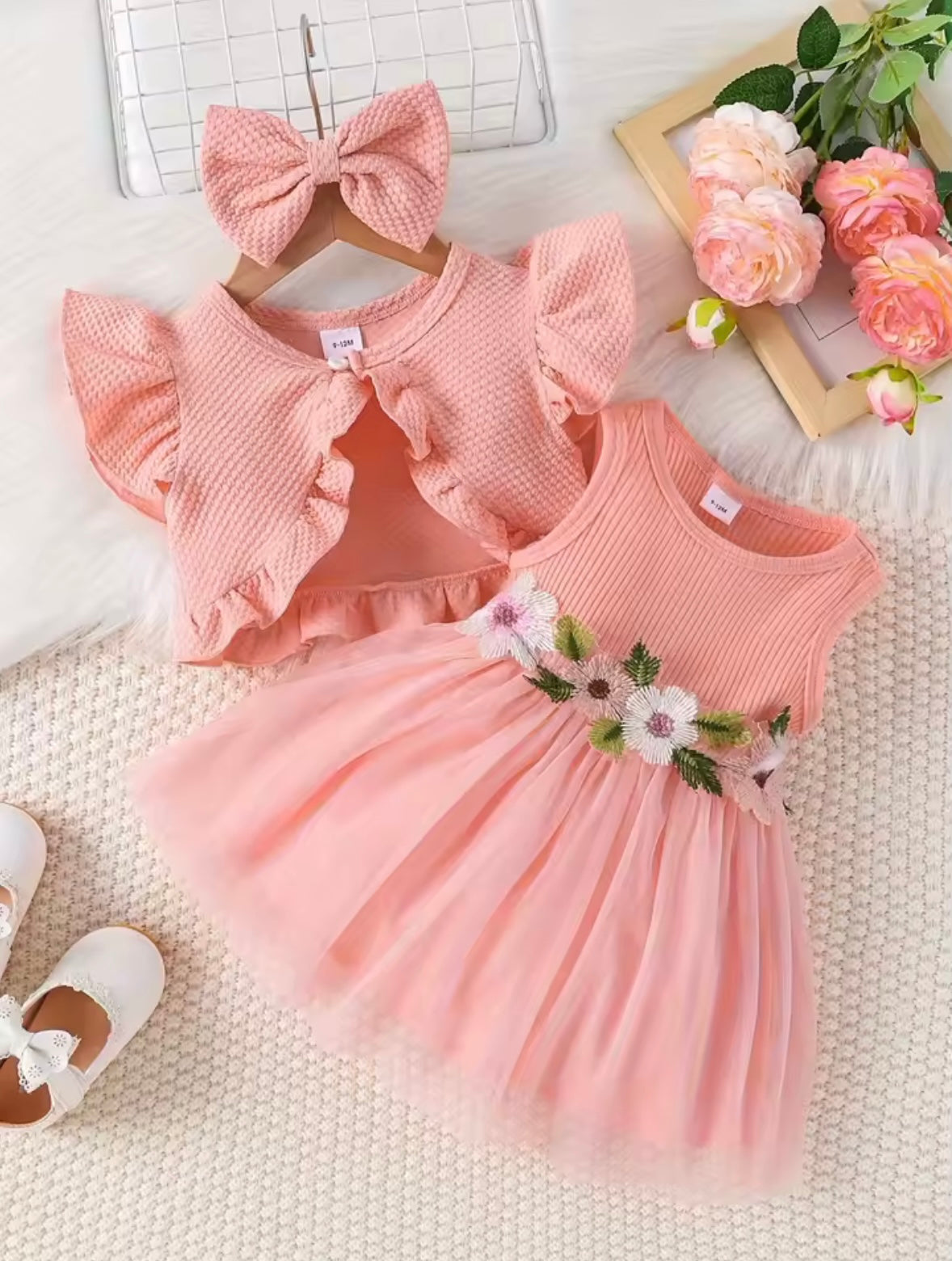 Pink baby dress with floral details, matching headband, and white shoes on a textured surface.