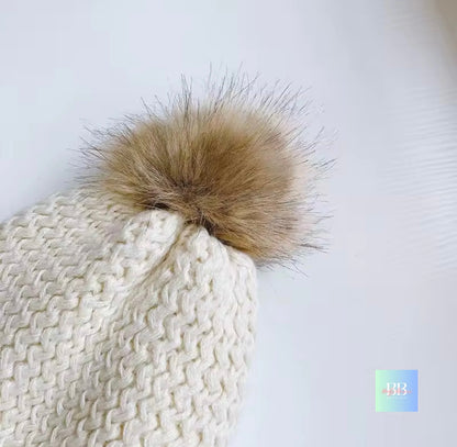 White knitted beanie with a large brown fur pom-pom on a light gray background