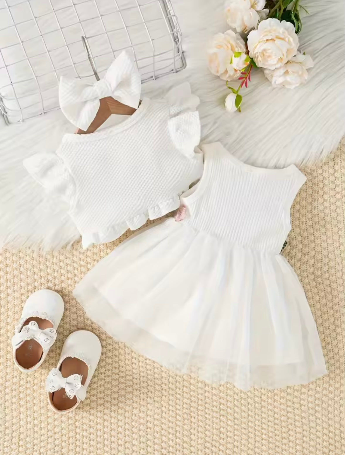 White baby dress with ruffled details, matching headband, and shoes on a textured surface with flowers.