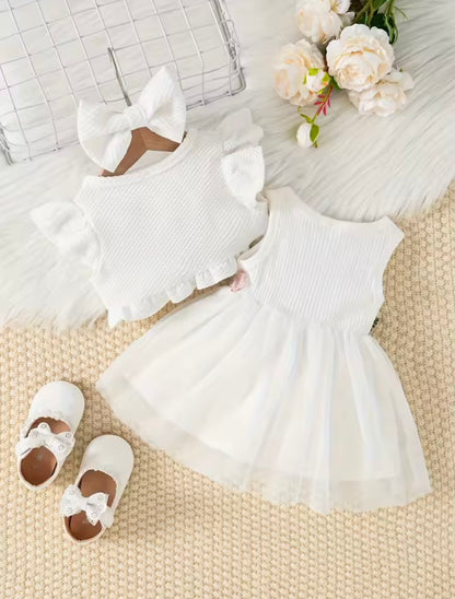White baby dress with ruffled details, matching headband, and shoes on a textured surface with flowers.
