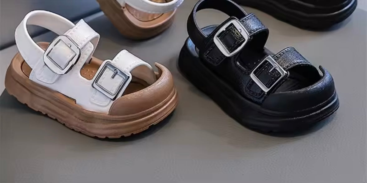 Pair of white sandals with brown soles and black sandals with silver buckles on a gray surface.