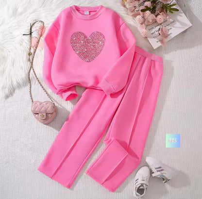 Pink sweatshirt with heart design and matching pants on a white surface with a small bag and shoes.
