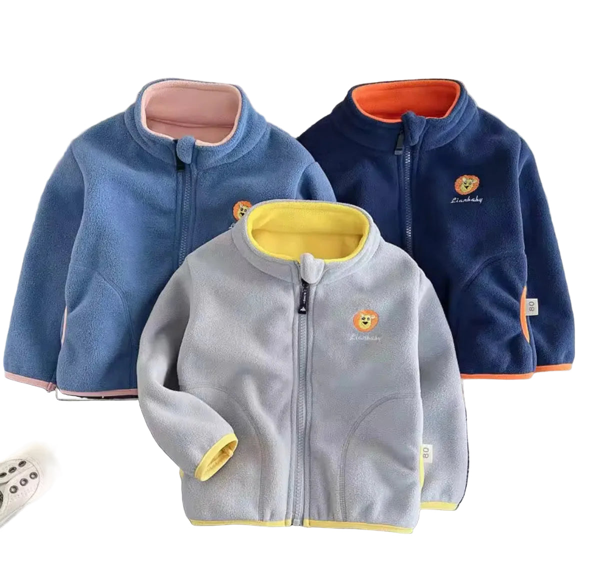 Three children's fleece jackets in blue, gray, and navy on a textured surface with shoes and a teddy bear.
