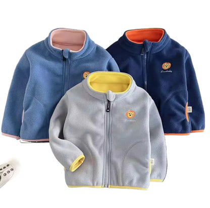 Three children's fleece jackets in blue, gray, and navy on a textured surface with shoes and a teddy bear.