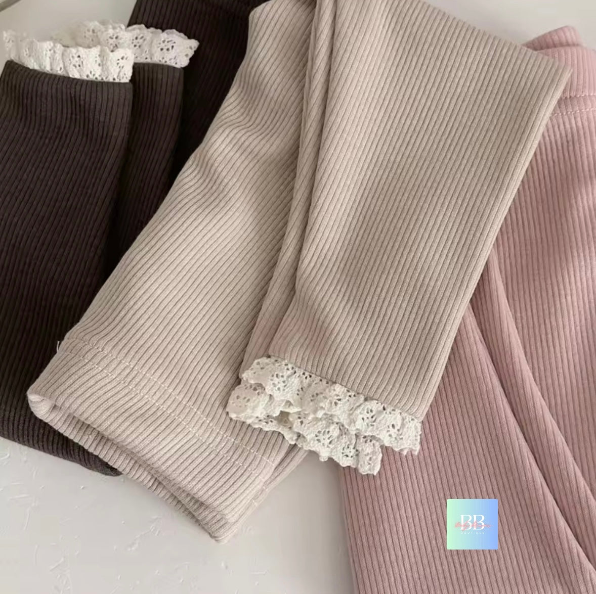 Three pairs of ribbed leggings with lace trim in black, beige, and pink on a light background.