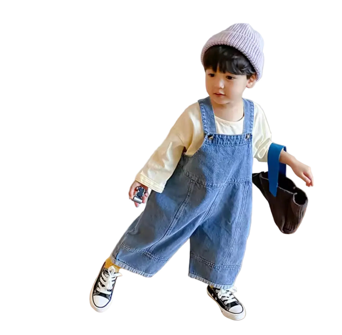 Child wearing a denim overalls and holding a small bag on a wooden floor.
