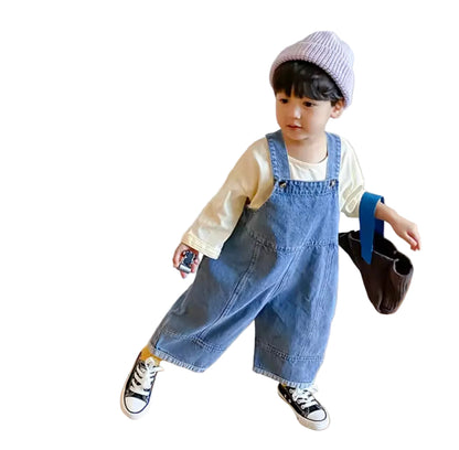 Child wearing a denim overalls and holding a small bag on a wooden floor.