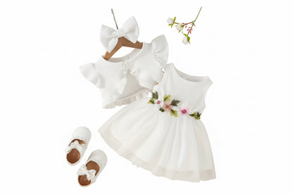White baby dress with floral details, white shoes, and headband on a textured surface with flowers.