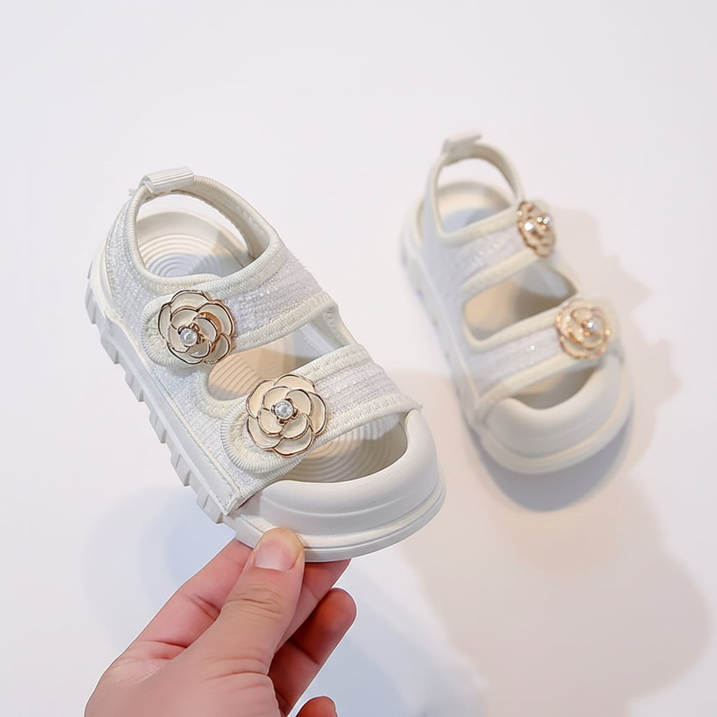 White children's sandals with floral decorations held by a hand on a neutral background