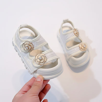 White children's sandals with floral decorations held by a hand on a neutral background
