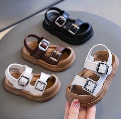 Children's sandals with buckle details on a gray surface, one sandal held by a hand.