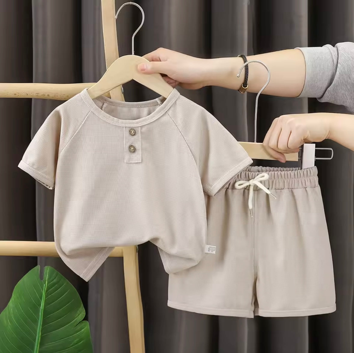 Children's beige outfit with shirt and shorts on hangers against a dark curtain background.