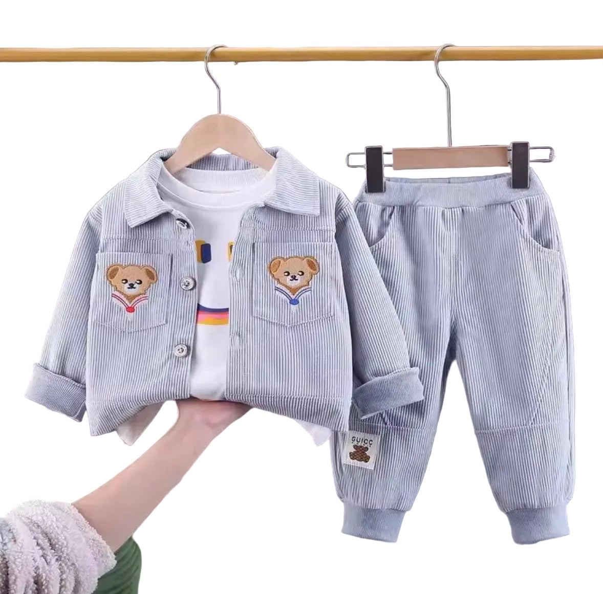 Children's clothing set with jacket, shirt, and pants on hangers against a gray curtain background.