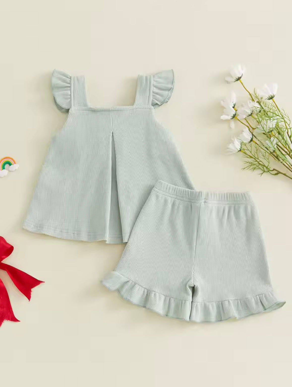 Light blue ruffled dress and shorts set on a beige background with flowers and a red bow.
